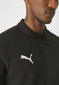 Čierna polo tričko s textúrovanou látkou, dvoma gombíkmi a bielym logom Puma na ľavej strane hrudníka.