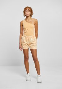 En-skulder, ferskenfarget tanktop med glatt stoff sammen med pastellfargede tie-dye shorts med elastisk liv. Hvite sneakers fullfører antrekket.