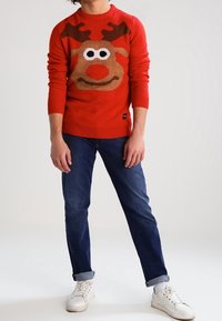 Personne portant un pull rouge avec un visage de renne, un jean bleu retroussé, des chaussettes blanches et des baskets blanches sur un fond uni.