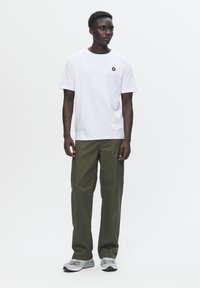 White cotton T-shirt with a black logo on the chest, paired with loose olive green pants and gray sneakers.