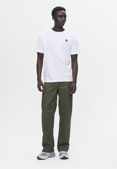 Weißes Baumwoll-T-Shirt mit einem schwarzen Logo auf der Brust, kombiniert mit lockeren olivgrünen Hosen und grauen Sneakers.