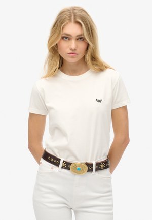 T-shirt bianca in cotone con scollo rotondo e maniche corte, caratterizzata da un piccolo logo nero. Abbinata a una cintura marrone e jeans bianchi.