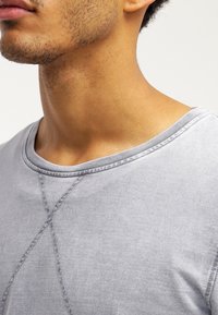 T-shirt in cotone grigio chiaro con scollatura rotonda, cuciture visibili e finitura texturizzata, caratterizzata da un sottile motivo a intreccio sul tessuto.