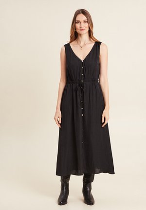 Zwart mouwloos maxi-jurk met een V-hals, voor knopen en een aanpasbare tailleband. G gedragen met zwarte laarzen, met een gladde textuur.