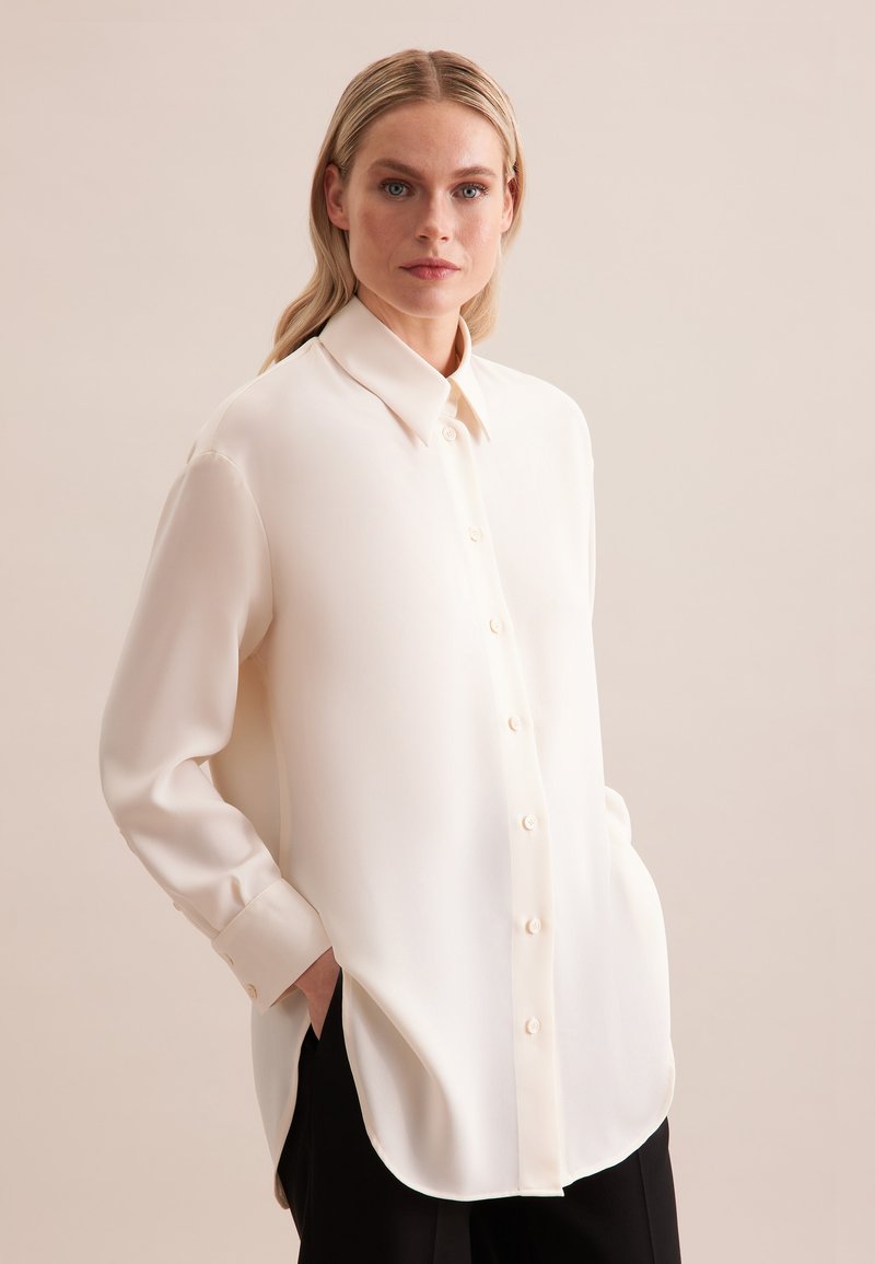 Femme aux cheveux blonds portant une chemise ample crème à manches longues et boutons, les mains dans les poches, sur un fond uni.