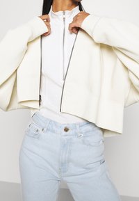 Chaqueta blanca recortada con mangas anchas sobre una camiseta blanca con cremallera, combinada con jeans de mezclilla azul claro con cierre de botón y bolsillos.