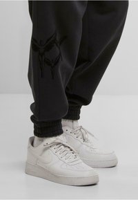 Hvide Nike sneakers med snørebånd, som bæres med sorte joggers, der har sorte englevinger grafikker på venstre ben.