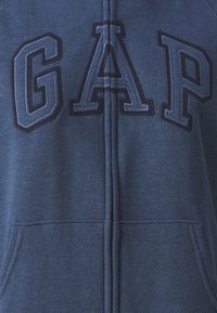 Sudadera con cremallera de color azul marino, con grandes letras bordadas "GAP" en un azul más oscuro, bolsillos frontales y suave textura de algodón.