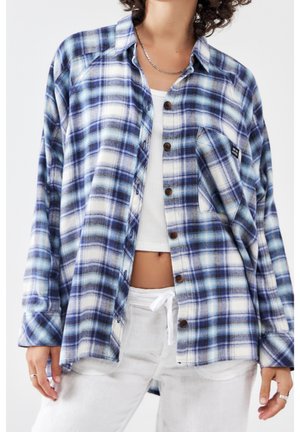 Persona que lleva una camisa de cuadros azul y blanca con botones sobre un top corto blanco y pantalones blancos con cordón, con un collar plateado.