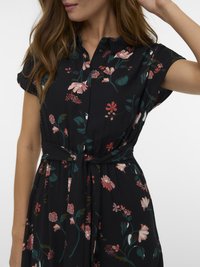 Γυναίκα που φοράει ένα μαύρο φλοράλ φόρεμα με κοντά μανίκια, γιακά και ζώνη στη μέση.