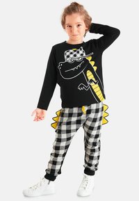 Schwarzes Langarmshirt mit Dinosaurier-Print und gelben Akzenten, kombiniert mit grau-schwarz karierten Hosen, die gelbe Stacheln und umgekrempelte Knöchel haben.