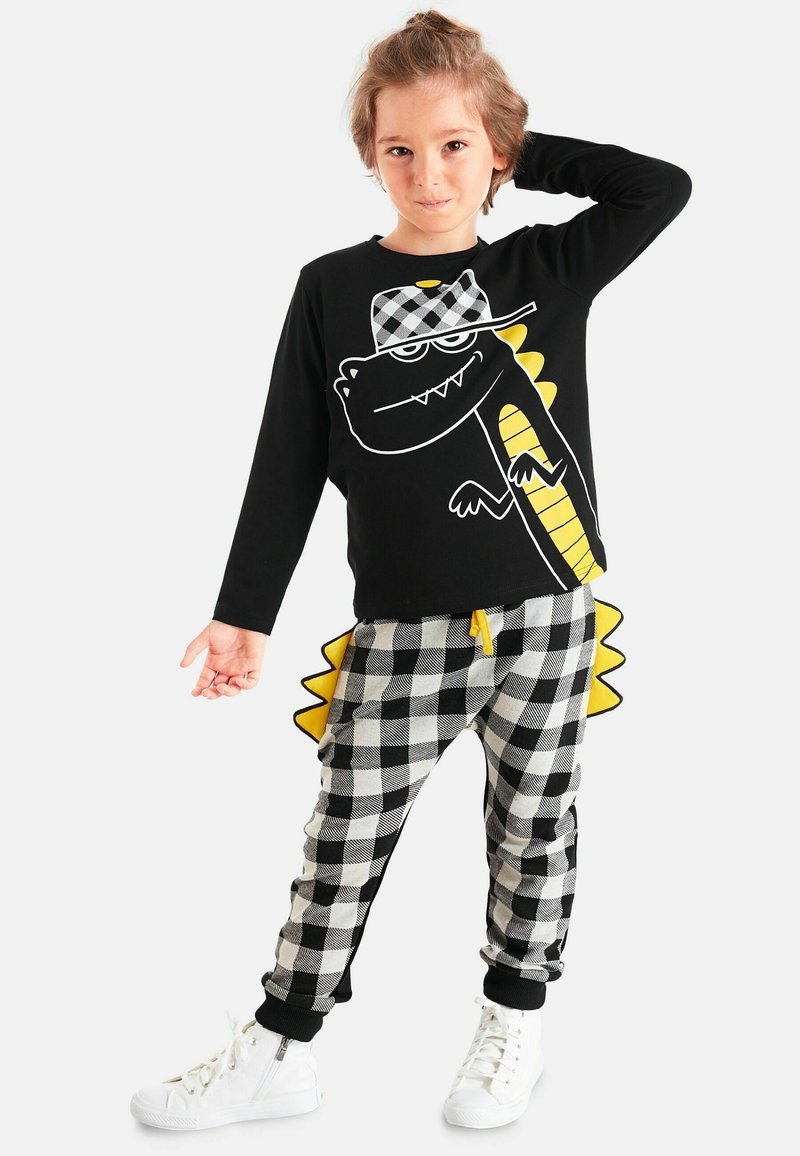 Schwarzes Langarmshirt mit Dinosaurier-Print und gelben Akzenten, kombiniert mit grau-schwarz karierten Hosen, die gelbe Stacheln und umgekrempelte Knöchel haben.