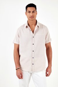 Mies, joka käyttää beigeä lyhythihaista napillista paitaa, jossa on käärityt hihansuut, ja valkoisia housuja, seisoo yksinkertaisen valkoisen taustan edessä.