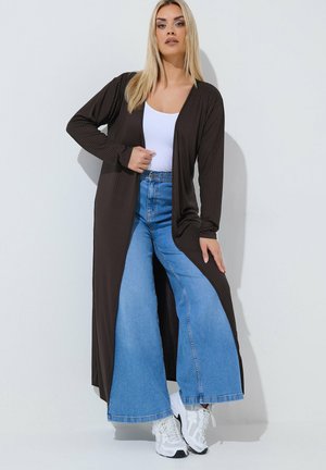 Kvinde iført en lang sort cardigan over en hvid top, brede blå jeans og hvide sneakers, stående mod en ensfarvet lys baggrund.