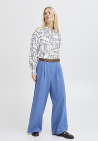 Camicia bianca con motivi geometrici neri, abbinata a pantaloni blue a gamba larga e una cintura intrecciata marrone, accentuata da scarpe nere.