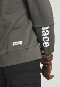 Sweat-shirt gris à manches longues, avec le texte blanc "MENACE" sur la manche droite et une étiquette cousue sur le côté gauche. Texture douce.