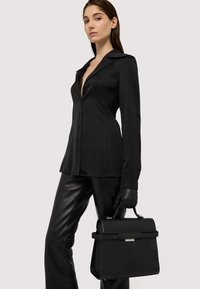 Sort satinknappet skjorte med dyb v-udskæring, parret med tætsiddende sorte læderbukser. Modellen holder en struktureret sort håndtaske.