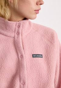Primo piano di una persona che indossa una giacca in pile rosa con bottoni a pressione e un logo Columbia nero sul petto.