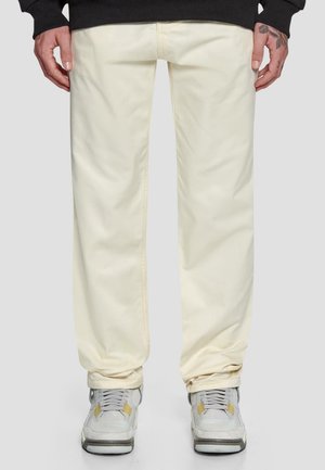 Beige Baumwollhosen mit lockerer Passform, die subtile Nähte aufweisen. Kombiniert mit grauen Sneakers mit gelben Akzenten.
