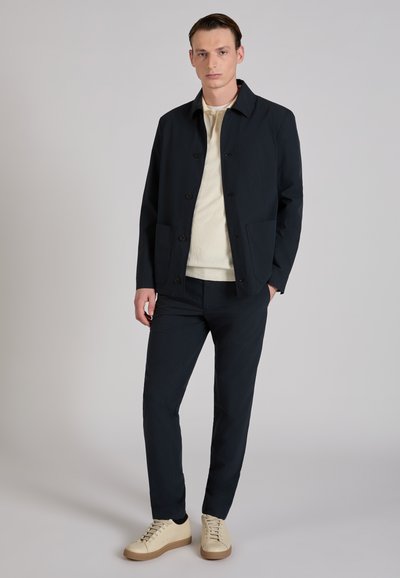 Homme portant une veste et un pantalon bleu marine, un pull crème et des baskets beige, debout avec une main dans la poche sur un fond gris clair.