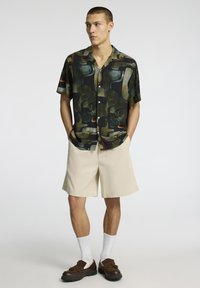 Kortärmad skjorta med knappar i ett abstrakt mönster av grönt och orange, ihop med beiga shorts. Bruna skor och vita strumpor kompletterar looken.