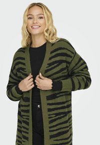 Olīvu zaļš kardigans ar melnām svītrām, ar brīvu piegriezumu, atvērtu priekšpusi un ribotām piedurknēm. Materiāls izskatās mīksts un ar tekstūru.