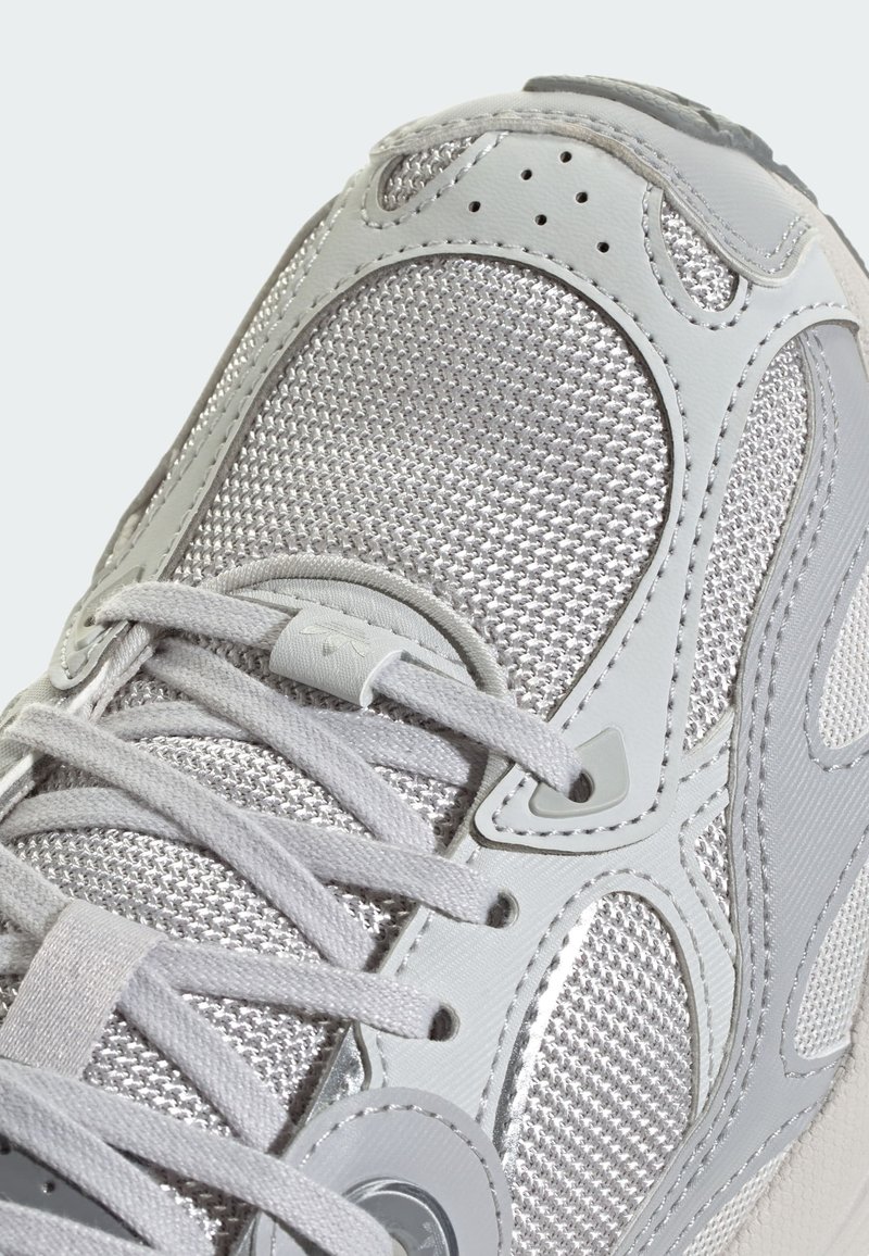 Zapatilla deportiva gris con parte superior de malla, superposiciones grises suaves, cordones redondos y suela texturizada. Acabada con sutiles detalles metálicos.