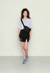 Modelo vestindo uma t-shirt oversized cinza-clara, saia preta com fendas laterais e ténis atléticos azuis, segurando uma pequena bolsa preta de ombro.