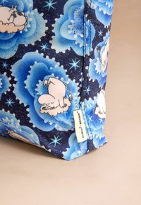 Pametna torba od pamuka s plavim cvjetnim uzorcima i bijelim Moomin likovima. Pozadina je tamnoplava s naglascima zvijezda. Oznaka je vidljiva sa strane.