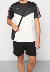 Man i svart och vit Nike sporttröja, svarta shorts och en svart axelväska mot en helt grå bakgrund.