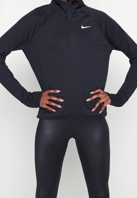 Zwarte, langmouwige Nike-top met een kwart rits, voorzien van een subtiel textuurpatroon. Gecombineerd met glanzende zwarte leggings.
