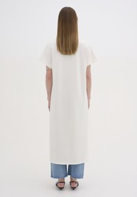 Kvinde med glat brunt hår iført en hvid kortærmet lang tunika over forkortede, slidte blå jeans og sorte hæle sandaler, stående med ryggen til.