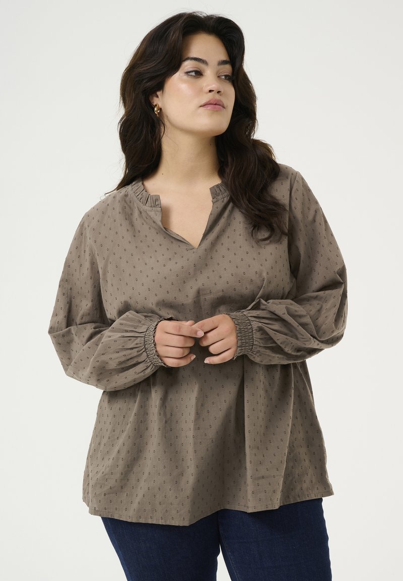 Langærmet, taupe bluse med en notch-udskæring, samlede manchetter og en subtil tekstur af små prikker. Løs pasform og blødt stof.