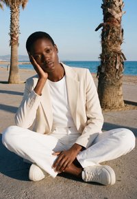 Jeune personne en blazer beige, chemise blanche et pantalon assise en tailleur sur le trottoir près des palmiers au bord de la mer, reposant la tête sur la main.