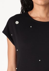 Black t-shirt with short sleeves, adorned with white pearl accents and a decorative flower brooch, featuring a smooth, soft texture.