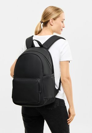 Rucsac sintetic negru cu curele căptușite, formă rotundă, buzunar frontal și un design minimalist. Finisaj texturat cu închideri prin fermoar.