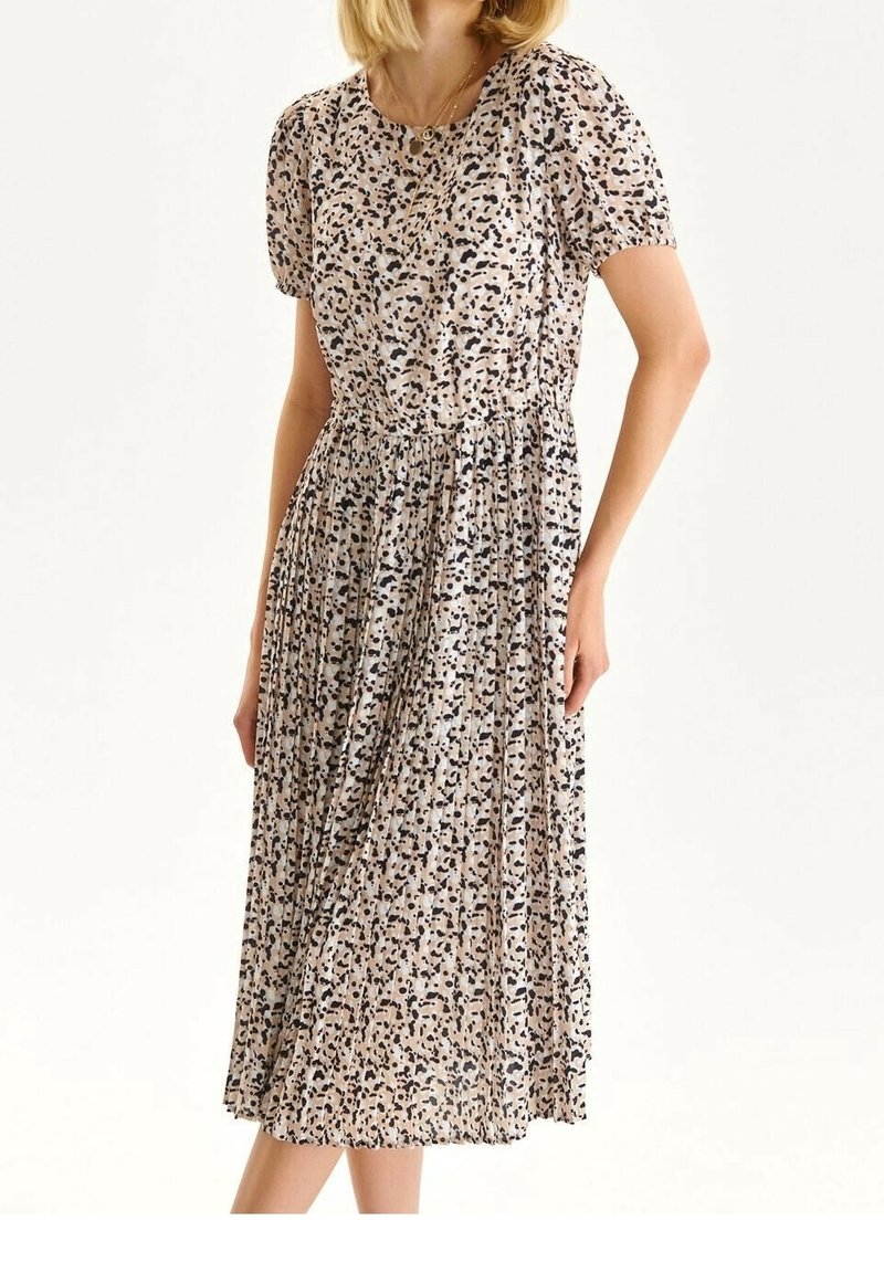 Femme portant une robe midi plissée beige avec des taches abstraites noires et brunes, des manches courtes bouffantes et un col rond.