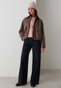 Bruine cropped jas over een lichtroze gebreide trui, gecombineerd met donkere wijde jeans en een gestructureerde roze beanie, afgemaakt met zwarte schoenen.