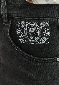 Čierne džínsy z denimu s čierno-bielou vzorovanou nášivkou na zadnom vrecku, ktorá obsahuje motívy paisley a podkovu.