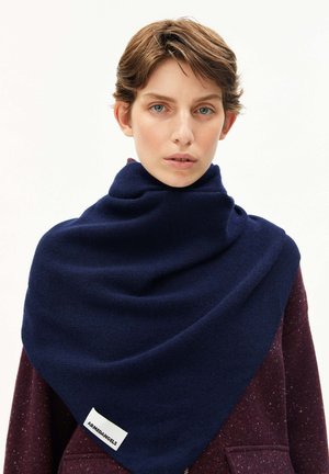 Person mit kurzem braunem Haar, die einen marineblauen dreieckigen Schal mit der Aufschrift "ARMEDANGELS" über einer kastanienbraun gesprenkelten Jacke trägt.