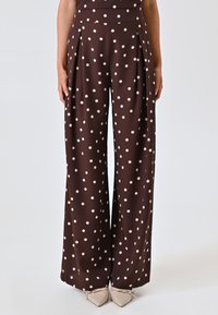 Bruine, wijde taillebroek met crèmekleurige stippen en plooien; gladde stof; hoge taille; geen zakken; gecombineerd met lichte puntige ballerina's.