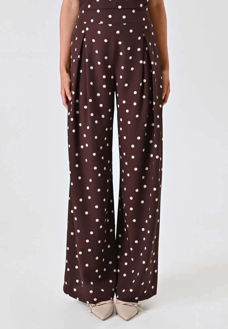 Bruine, wijde taillebroek met crèmekleurige stippen en plooien; gladde stof; hoge taille; geen zakken; gecombineerd met lichte puntige ballerina's.
