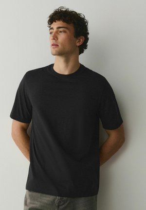 Fiatal férfi göndör barna hajjal, fekete rövid ujjú pólóban, világosszürke falnak támaszkodva, oldalra néz.