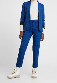 Blå rutig kostym med svarta accenter, utrustad med en åtsittande kavaj och smalare byxor, kombinerat med stiliga vita sneakers och en krämfärgad polo.