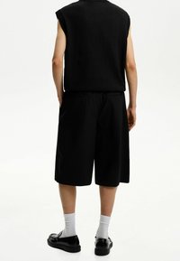 Svart ärmlös topp med en texturerad yta, kombinerad med svarta knälånga shorts, bärs med vita strumpor och svarta skor, sedd bakifrån.