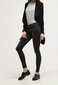 Cardigan lungo nero sopra una maglietta grigia, abbinato a jeans scuri attillati e scarpe nere lucide. Il tessuto è liscio con una vestibilità sartoriale.
