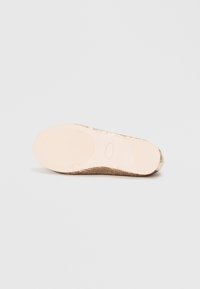 Flat-soled fottøy med en lys beige gummibase, med et teksturert gyllent stoffoverdel og et gjennomsiktig element nær hælen.