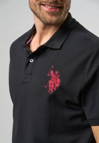 Černé polo tričko s texturovanou látkou, s červeně vyšitým logem na levé straně hrudi a jemným vzorovaným límcem v červené a bílé barvě.