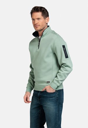 Lichtgroene zip-up sweatshirt met een hoge kraag, voorzien van een ritszak op de linker mouw en geribbelde manchetten. Gecombineerd met donkere jeans.