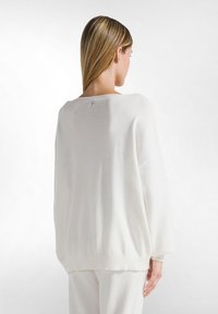 Weißer, übergroßer Strickpullover mit weitem Ausschnitt, überschnittenen Schultern und geripptem Saum. Weiche Textur mit einem dezenten Etikett auf der Rückseite.